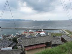 A scenic view overlooks a harbor with two red-and-white ferries, a town with traditional tiled roofs, and a calm sea with distant islands under a cloudy sky, framed by power lines.