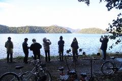 A group of tourists on motorcycles and bicycles observe a serene lake and forested hills on a clear day.