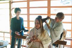 A bride in traditional attire is having her hair styled by a woman while another person stands nearby.