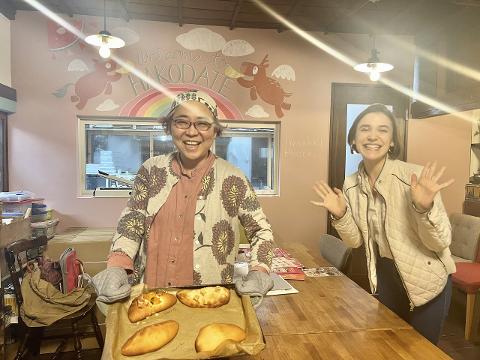 Two women, one holding a tray of freshly baked piroshki, smile in a charming Hakodate bakery with a whimsical mural and handwritten menu.