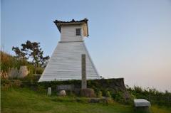 A white, pyramidal structure with a small window and tiled roof stands on a grassy hill, with a stone monument in front of it, under a clear sky.