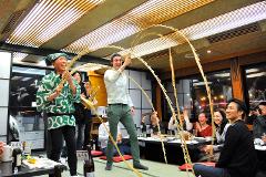 A group of people are enjoying a celebration or performance, with two men holding arched sticks and others clapping and taking photos at tables in a restaurant.
