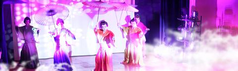 A group of people in traditional Japanese clothing are performing on stage with umbrellas in their hands, illuminated by pink and white lighting and fog.