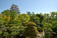 A multi-tiered Japanese castle rises above lush green trees, creating a picturesque scene under a clear blue sky.