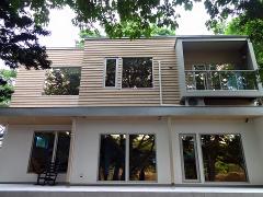 A contemporary two-story house with wood siding on the upper level, large reflective windows, and a small balcony on the right side, surrounded by trees.