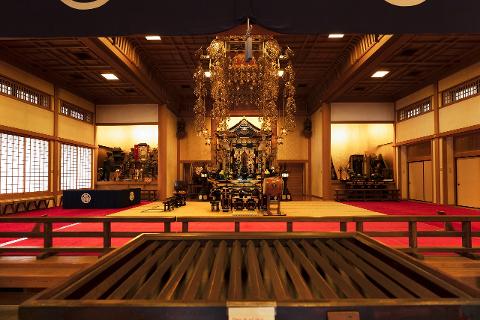 The ornate interior of a Japanese temple features a gilded altar, traditional shoji screens, and a vast hall with red carpeting.