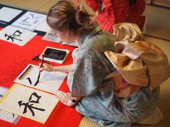 Here's a one-sentence description of the image:A woman in a traditional Japanese kimono practices calligraphy on a red surface with sample characters displayed.