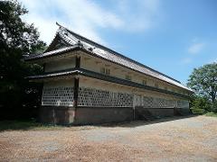 A long, traditional Japanese building with a tiled roof stands under a clear blue sky, surrounded by trees and dry grass.