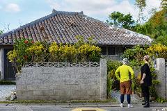 Two tourists stand admiring a traditional Okinawan home with a tiled roof and lush garden, embodying the wellness culture and longevity celebrated in Shioya Bay.