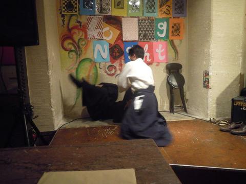 Two individuals are engaged in a martial arts demonstration on a stage decorated with colorful artwork and lettering spelling "NIGHT", with a stool and equipment visible in the background.