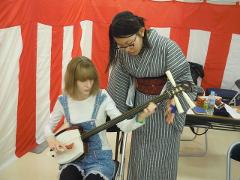 To kvinder spiller på en shamisen, en trestrenget traditionel japansk lut. Den ene har en stribet kimono på og underviser den anden, som har overalls på. De står foran en rød-hvidstribet baggrund.