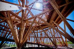 The image shows a wooden architectural structure against a bright blue sky with a sunburst effect, possibly at an art festival.