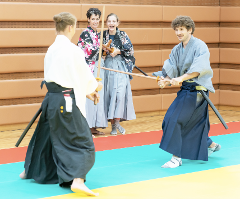 Dvaja ľudia v tradičnom japonskom oblečení si precvičujú šermiarsky štýl s drevenými mečmi, zatiaľ čo dve ženy ich s úsmevom sledujú.