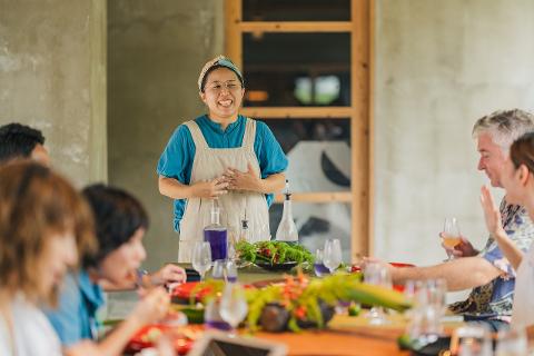 A smiling chef in an apron stands at the head of a dining table, addressing a group of people enjoying lunch with colorful drinks and dishes.