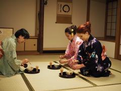Three women in kimonos sit on tatami mats, participating in a traditional Japanese tea ceremony, each with their own set of tea utensils.