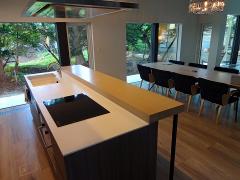 A modern kitchen island with a cooktop and sink sits adjacent to a dining table in a bright, open space with large windows overlooking lush greenery.