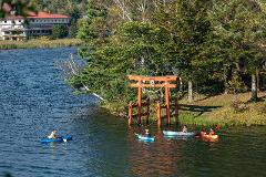 Visit the Spring Water Flowing into Lake Shirakaba and Experience the Origins of Our Connection with Nature – Private Guided Canoe Tour Kayakers paddle on a serene lake in front of a Japanese torii gate with a hotel visible in the background.