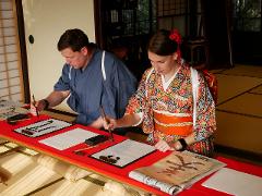 Two people dressed in traditional Japanese clothing are practicing calligraphy with brushes and ink on white paper at a red table.