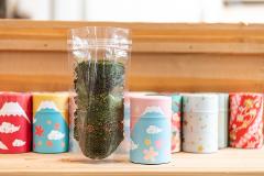 A clear plastic bag of green tea leaves sits in front of a row of decorative Japanese tea tins featuring Mt. Fuji and cherry blossoms.