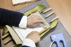A craftsman's hands are shown working on a traditional tatami mat, securing fabric with staples near a blue staple gun and wooden tools.