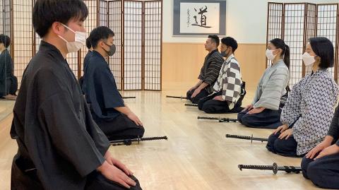 Un groupe de personnes en vêtements traditionnels japonais sont agenouillées en rang sur un plancher en bois avec des épées devant elles.