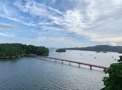 A bright red bridge spans a calm bay dotted with boats, connecting lush green islands under a partly cloudy sky.