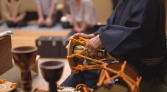 Mukojima Geisha Experience: Training Session & Ozashiki Banquet at a Ryotei Restaurant A performer in traditional Japanese attire plays a small drum with orange cords, surrounded by musical instruments and other seated individuals.
