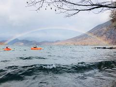 Two people kayak on a lake under a rainbow as mountains rise in the background.