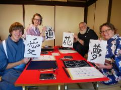 Four people wearing traditional Japanese clothing pose behind a red table, each holding up sheets of paper with calligraphy.
