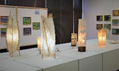 A display of various handmade paper lamps illuminated with warm light sits on a table in front of artwork.