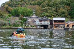A person kayaks on a calm lake in front of a village nestled at the base of a forested hill.