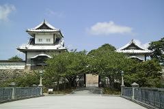 The image shows the entrance gate and a turret of a traditional Japanese castle, surrounded by lush green trees, under a clear blue sky with scattered clouds.