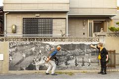 A man and a woman admire a black-and-white mural of a traditional Japanese festival on a stone wall in front of a building.