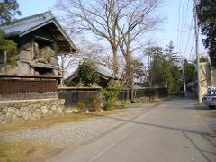 A silver car is parked on the side of a paved road in a rural area with traditional Japanese buildings and trees.