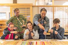 A group of people are gathered around a table, engaged in a craft activity, with a man in traditional Japanese attire demonstrating how to tie mizuhiki knots.