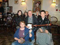 En gruppe mennesker poserer sammen, en mand forrest spiller en shamisen, en anden holder en, med et maleri af en geisha i baggrunden.