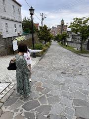 A woman in a green dress and a guide stand on a cobblestone street in Hakodate, with the guide pointing towards a building in the distance.