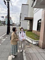Two women stand on a sidewalk, one in a patterned dress and jacket, the other in a white jacket and mask, with buildings and a street behind them.