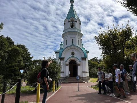A group of tourists is admiring the Hakodate Orthodox Church on a partly cloudy day.
