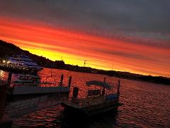 A picturesque view of a harbor at sunset, with vibrant orange and yellow hues painting the sky and reflecting on the water, alongside boats and a pier.