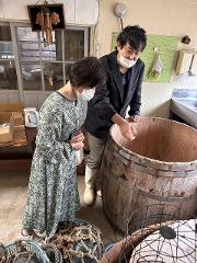 A man and woman in masks look inside a large wooden barrel, surrounded by glass buoys and other items in a room with a window and various objects hanging on the walls.