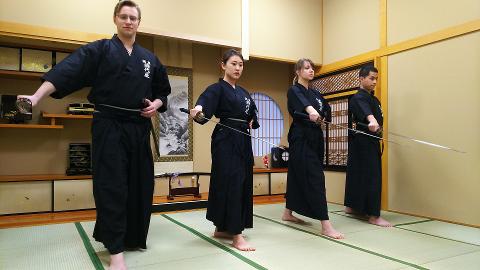 Four people in traditional Japanese attire practice iaido with swords in a tatami room.