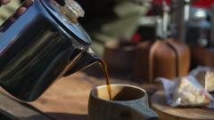 Coffee is poured from a stainless steel pot into a wooden mug, with a pastry and camping equipment visible in the background.