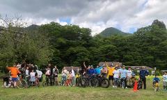 A large group of people with bicycles in a grassy area with trees and mountains in the background.