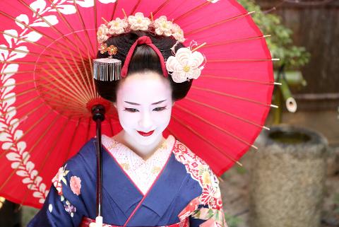A woman in traditional Japanese geisha attire holds a red umbrella, her face painted white with red lips and her hair adorned with flowers and ornaments.