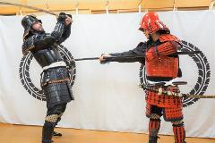 Two men in samurai armor face each other with swords drawn in a studio.