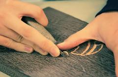 A person's hands carve a design into a piece of dark wood with a tool, creating wood shavings.