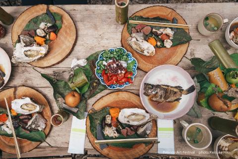 A bird's-eye view of a rustic outdoor table laden with a variety of Japanese dishes, including oysters, grilled fish, and seafood soup, presented on wooden slices and banana leaves.