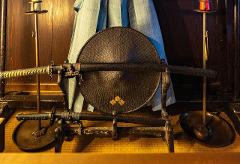 Two samurai swords are displayed on a stand in front of a woven hat, with decorative candles and a light blue garment in the background.