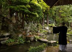 A monk stands with arms outstretched beside a pond in a serene Japanese garden.